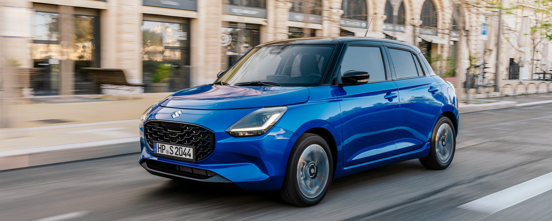 Blauer Suzuki Swift fährt dynamisch durch eine Straße in städtischer Umgebung