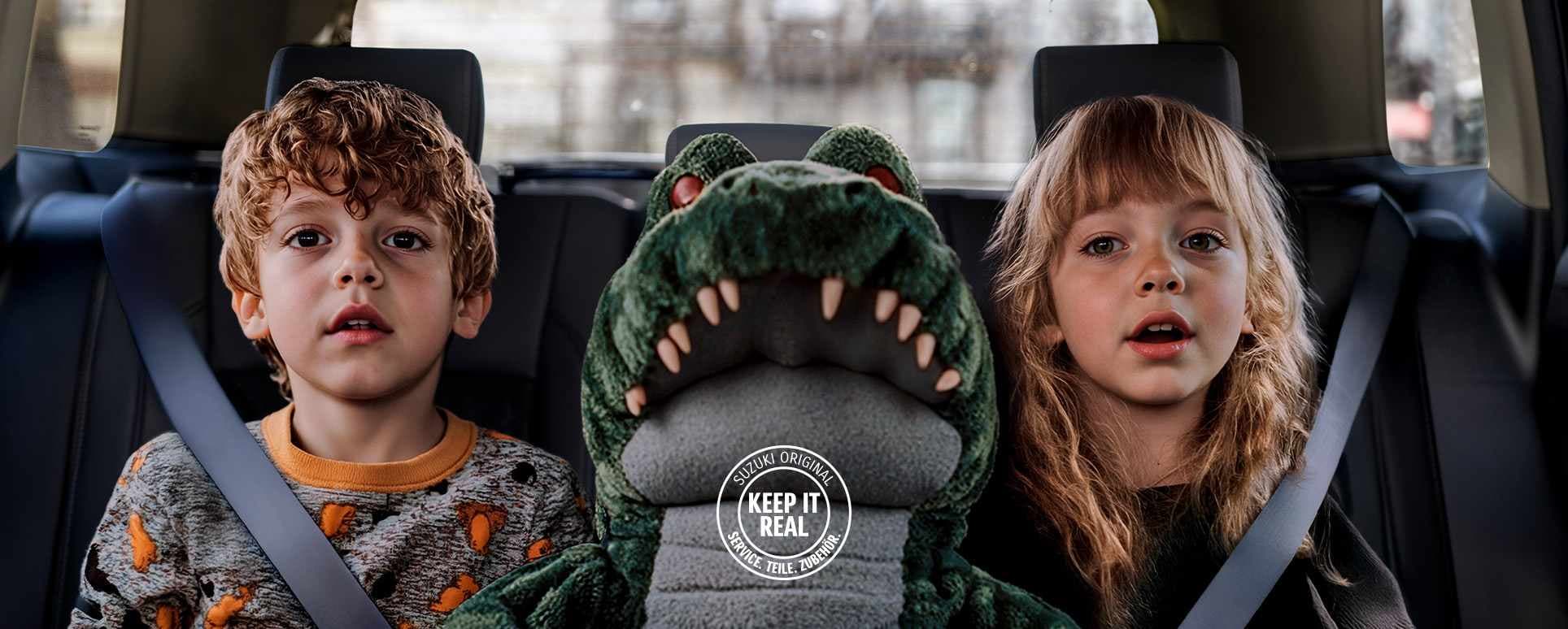 Zwei kleine Kinder sitzen angeschnallt auf einer Autorückbank, zwischen sich ein großes Plüschkrokodil, im unteren Bereich ist das Logo „Keep it real“ in weiß zu sehen