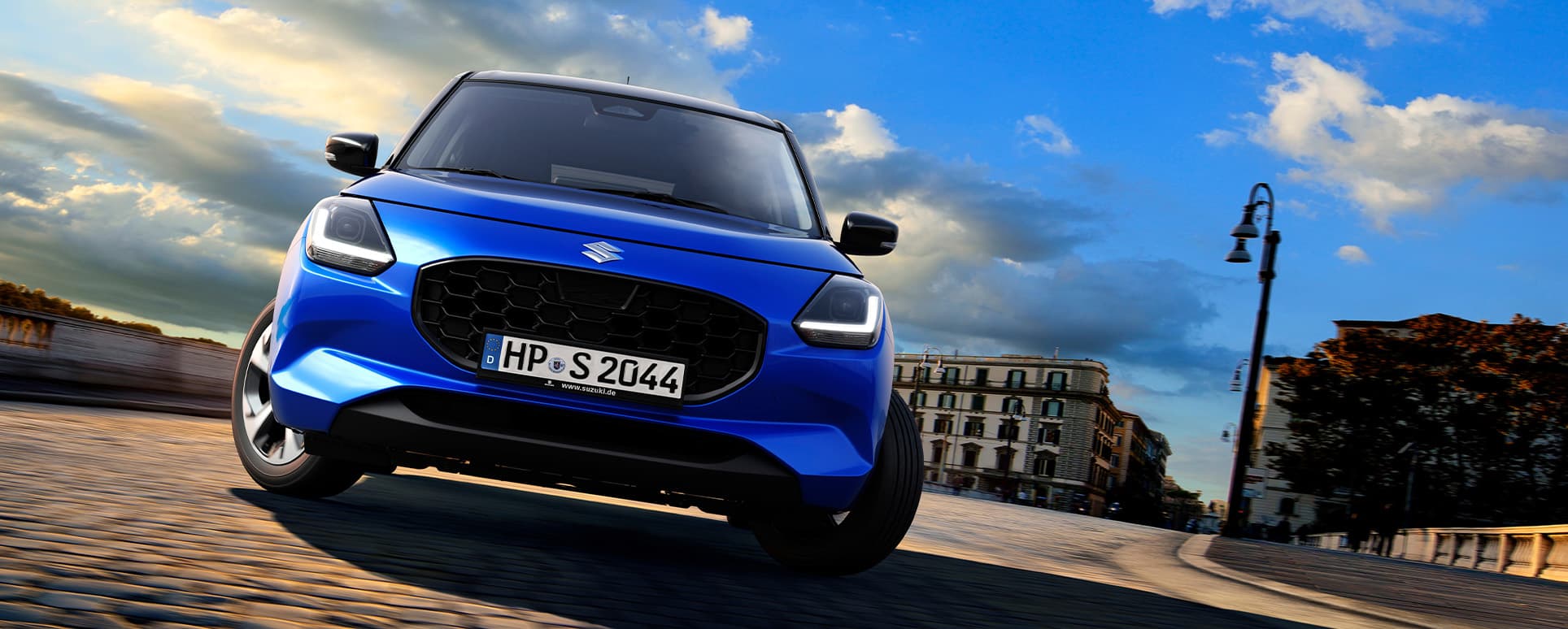 Ein blauer Suzuki Swift fährt in Frontalansicht um eine Kurve in städtischer Umgebung