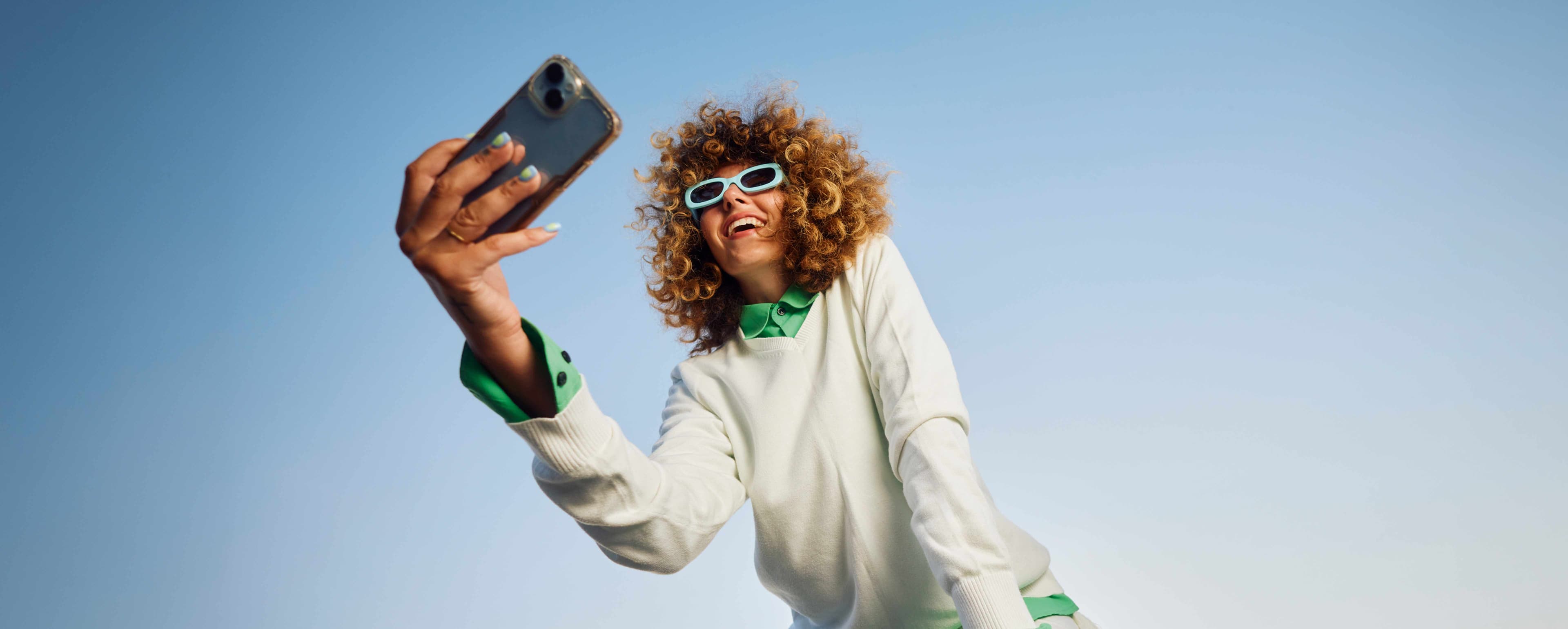 Frau in mindgrünem Pullover mit Sonnenbrille schaut auf ihr Smartphone und lacht