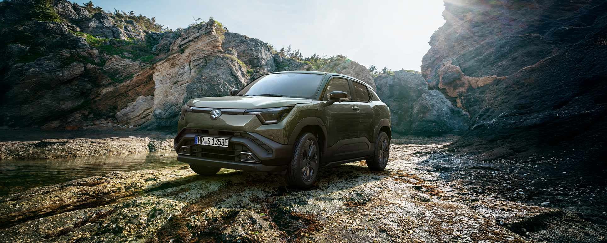 Der e VITARA in dunkelgrün vor einer felsigen Outdoor-Kulisse neben einem kleinen Gewässer.