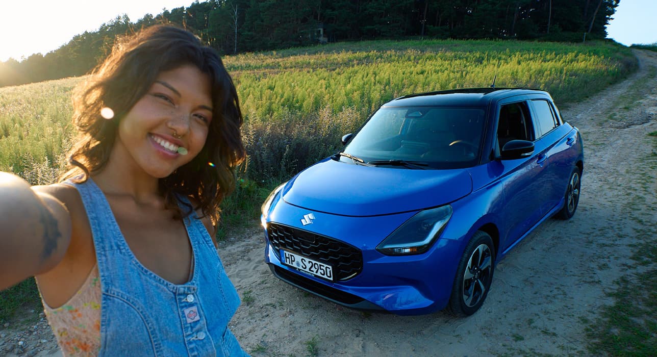 Frau macht ein Selfie auf einem Feldweg mit einem Suzuki Mild-Hybrid im Hintergrund