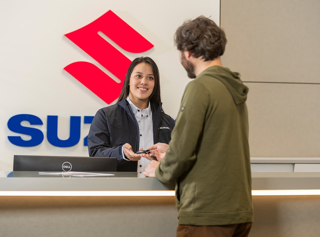Eine Frau an einem Empfangsschalter mit großem Suzuki-Schriftzug überreicht einem Mann im Kapuzenpullover einen Autoschlüssel