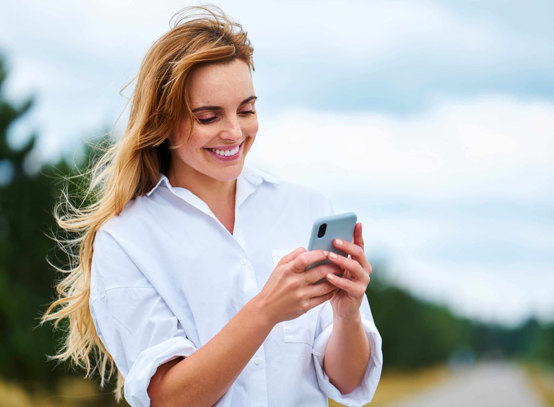 Frau in einem weißen Hemd steht am Straßenrand und schaut auf ihr Smartphone