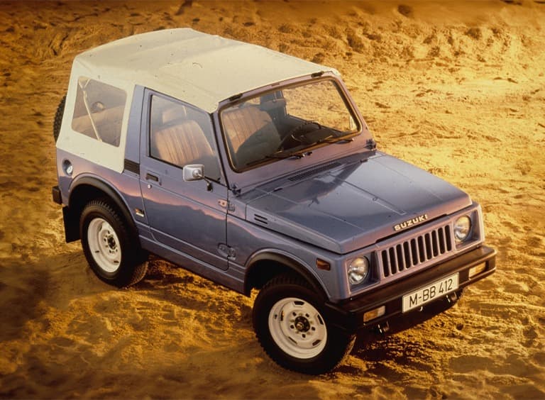 Abbildung zeigt von schräg oben einen blauen Jimny mit einem weißen Dach, der im Sand parkt