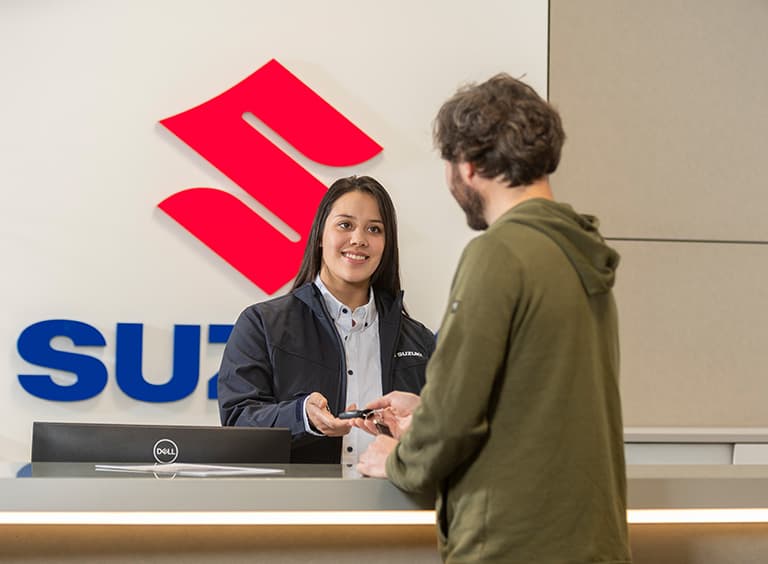Eine Frau an einem Empfangsschalter mit großem Suzuki-Schriftzug überreicht einem Mann im Kapuzenpullover einen Autoschlüssel