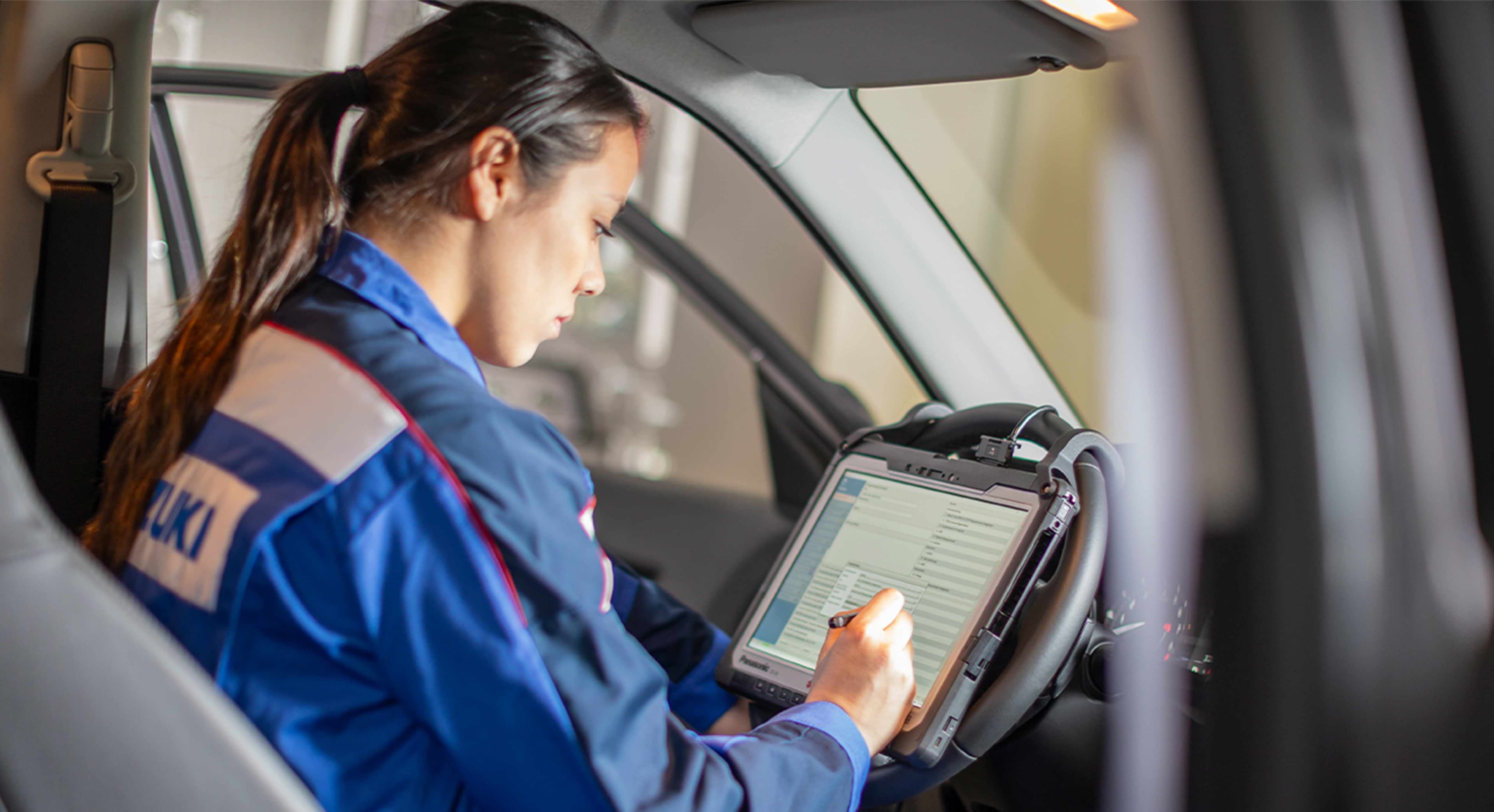 Eine Suzuki Mitarbeiterin sitzt in einem Suzuki Overall in einem Auto. Sie arbeitet an einer auf einem Tablett dargestellten Checkliste.