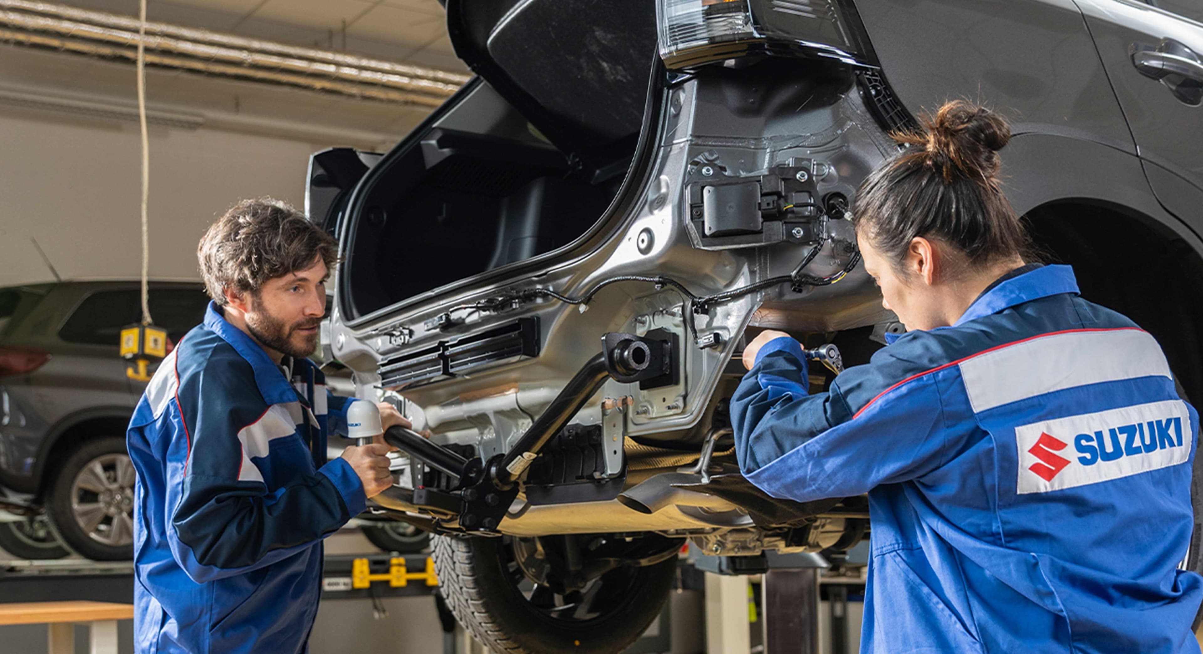 Ein Mann und eine Frau im blauen Suzuki-Arbeitsoutfit arbeiten an einem Auto, das mit der Hebebühne hochgefahren wurde
