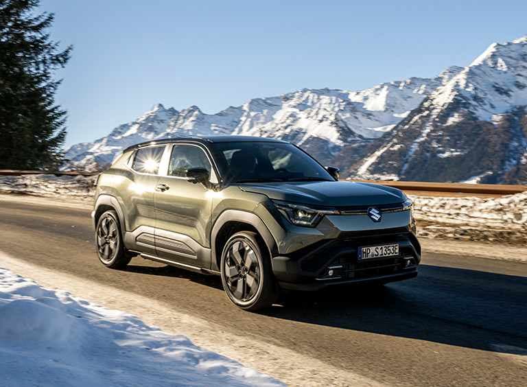 Suzuki e VITARA fährt auf einer Landstraße mit winterlicher und verschneiter Bergkulisse im Hintergrund.
