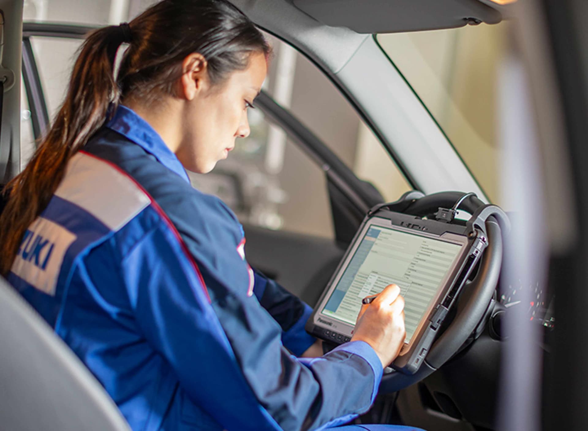 Eine Suzuki Mitarbeiterin sitzt in einem Suzuki Overall in einem Auto. Sie arbeitet an einer auf einem Tablett dargestellten Checkliste.