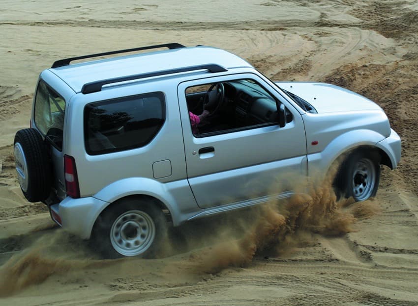 Abbildung zeigt einen weißen Jimny, der eine Düne hinauffährt