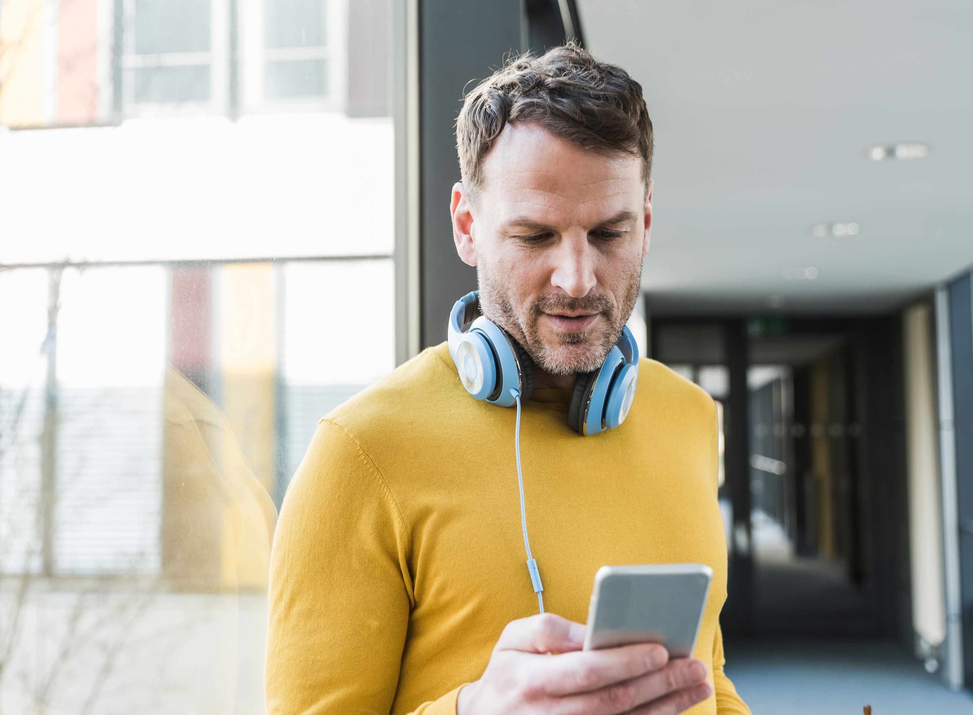 Mann in einem gelben Pullover und mit Kopfhörern um den Hals schaut auf sein Smartphone