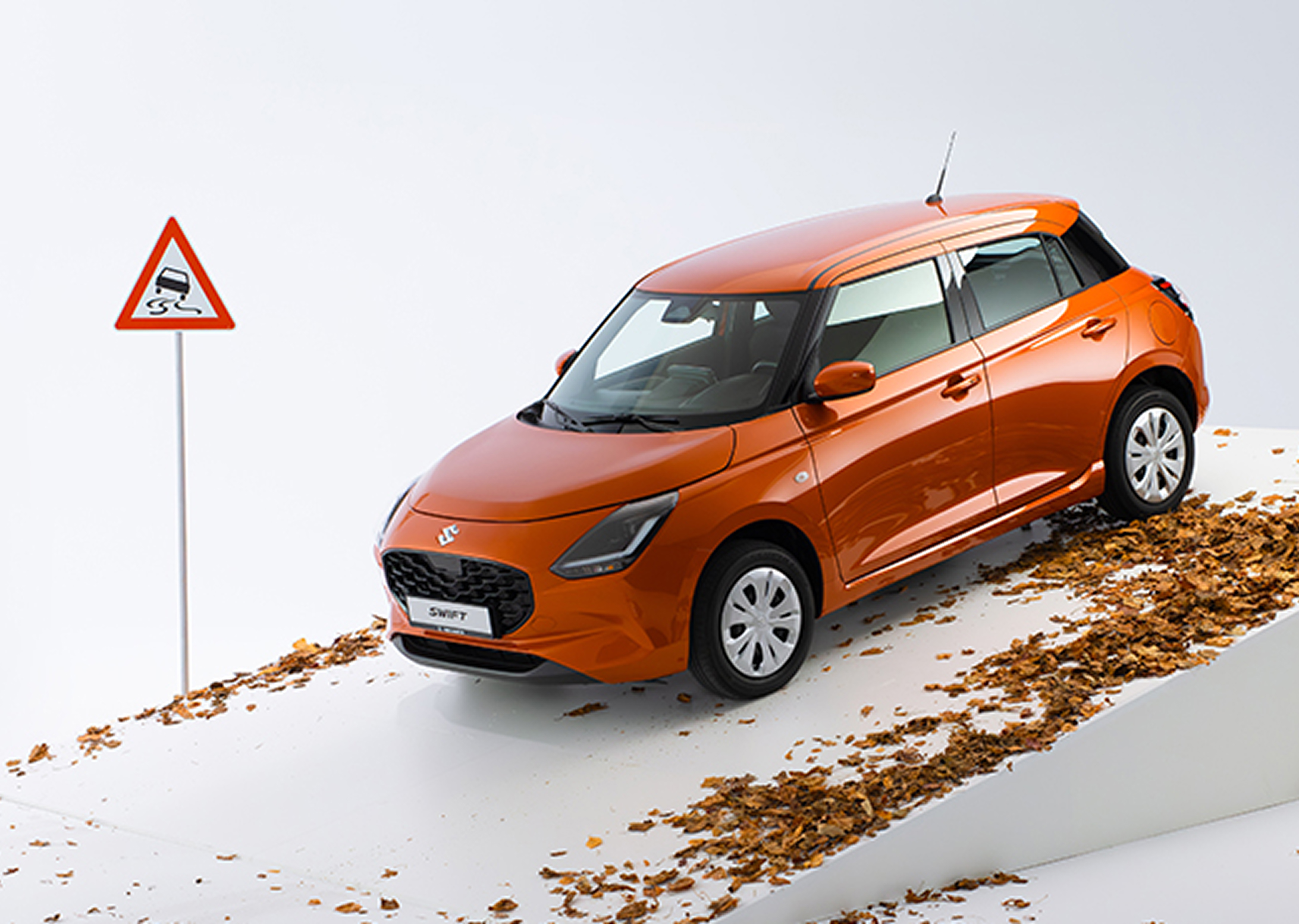 Der Suzuki Swift in orange steht auf einer weißen Rampe mitsamt Laub, daneben ein Verkehrswarnschild für Rutschgefahr. Eine lächelnde junge Frau in legerer Kleidung lehnt an der Tür auf der Fahrerseite.