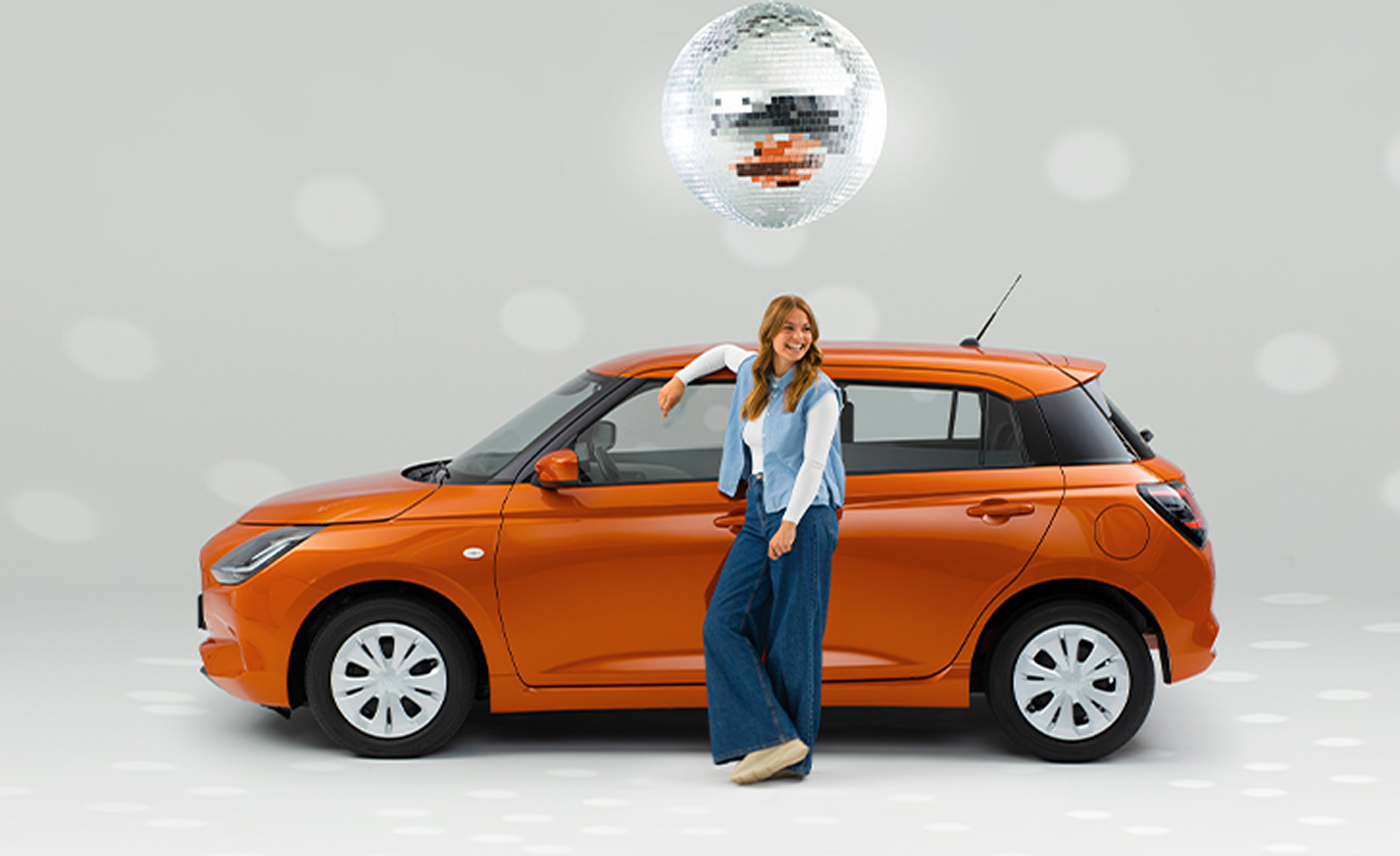 Eine junge Frau in legerer Kleidung steht lachend vor einem seitlich geparkten Suzuki Swift in orange, über ihr wirft eine Discokugel Reflektionen auf den weißen Hintergrund.