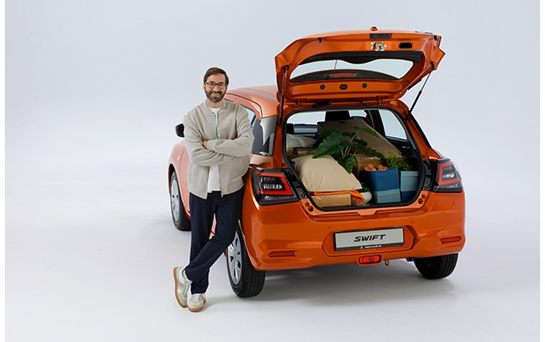 Ein Mann in legerer Kleidung mit Bart und Brille steht vor einem Suzuki Swift in orange mit geöffnetem, beladenem Kofferraum.