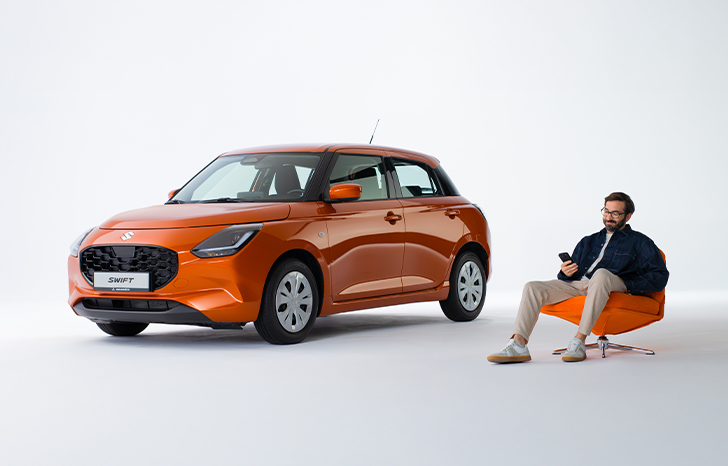 Ein Mann in legerer Kleidung mit Bart und Brille sitzt in einem orangefarbenen Sessel neben einem Suzuki Swift in Orange, er blickt lächelnd auf das Display eines Smartphones in seiner Hand.