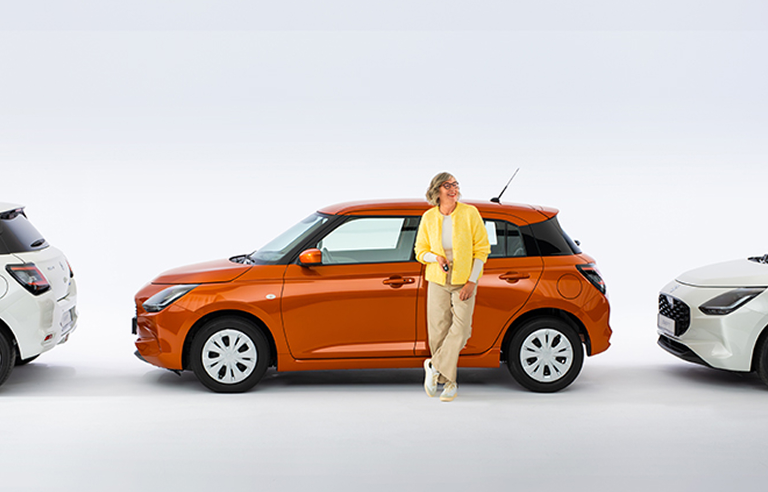 Eine lachende Best-Agerin in legerer Kleidung mit Brille steht vor einem seitlich parkenden Suzuki Swift in Orange, der Hintergrund ist weiß und neutral.