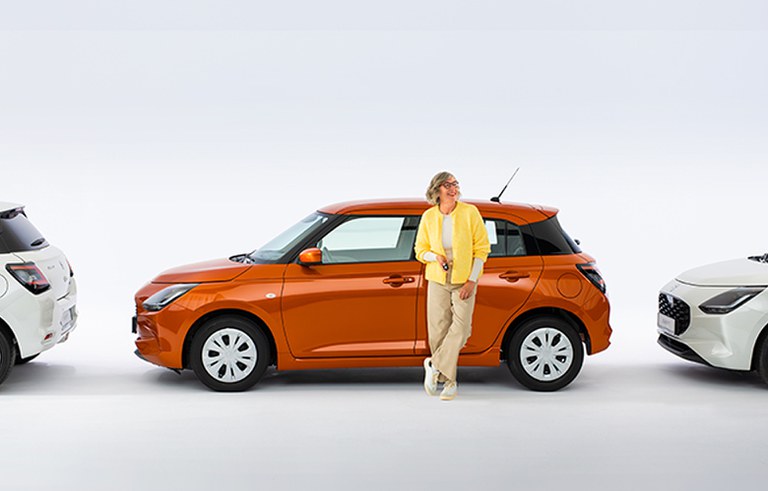 Eine lachende Best-Agerin in legerer Kleidung mit Brille steht vor einem seitlich parkenden Suzuki Swift in Orange, der Hintergrund ist weiß und neutral.