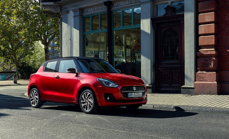Seitliche Aufnahme eines Suzuki Swift in Burning Red Pearl Metallic, vor einem Cafe parkend. Suzuki Swift Hybrid in Burning Red Pearl Metallic parkt vor einem Gebäude an der Straßenecke.