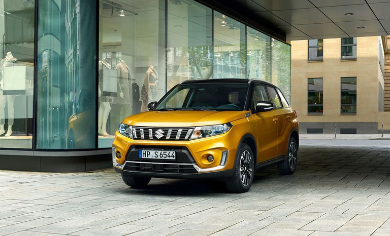 Suzuki Vitara Hybrid in Solar Yellow Pearl Metallic steht vor einem Modegeschäft. Suzuki Vitara Hybrid in Solar Yellow Pearl Metallic steht vor einem Modegeschäft.