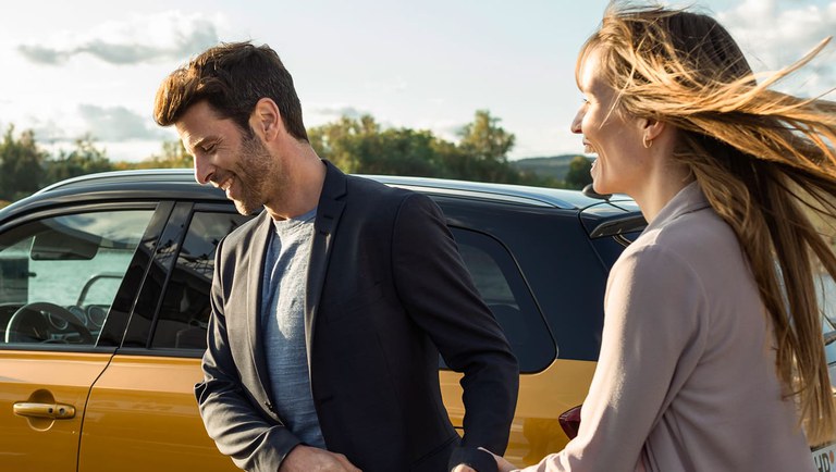 Eine Frau und ein Mann laufen von rechts nach links, vor einem Suzuki Vitara Hybrid in Solar Yellow Pearl Metallic. Eine Frau und ein Mann laufen von rechts nach links, vor einem Suzuki Vitara Hybrid in Solar Yellow Pearl Metallic.