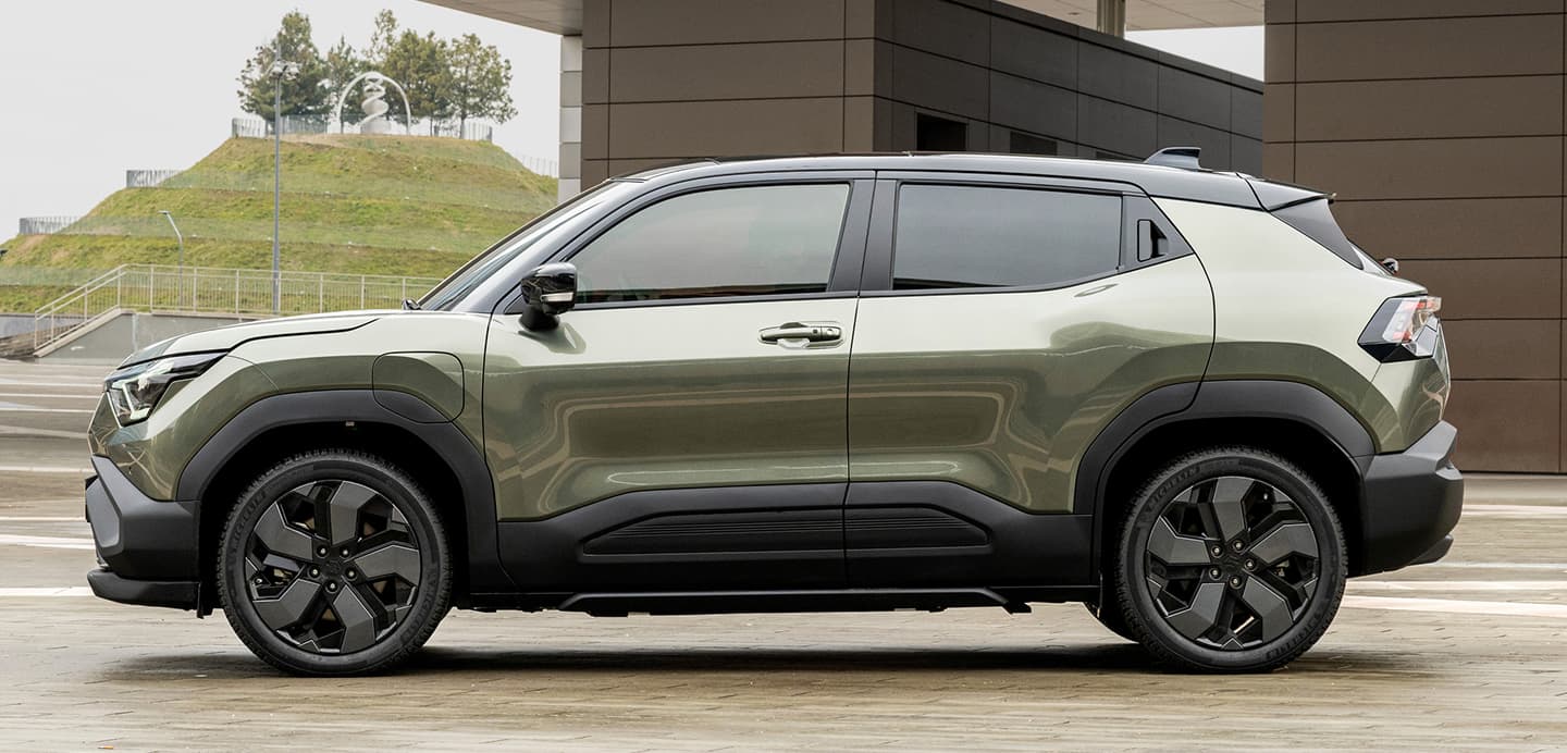 Der e VITARA in dunkelgrün parkt vor einem Gebäude neben einem grasbewachsenen Abhang