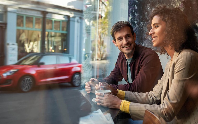 Paar in Kaffee mit Suzuki Swift im Hintergrund Mann und Frau sitzen in einem Café und schauen raus auf einen Suzuki Swift in Red Pearl Metallic.