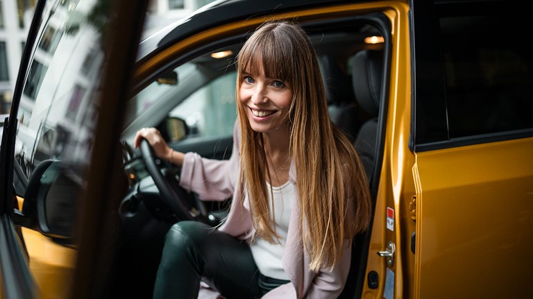 Frau mit langen braunen Haaren steigt aus dem Suzuki Vitara Hybrid in Solar Yellow Pearl Metallic und grinst in die Kamera.