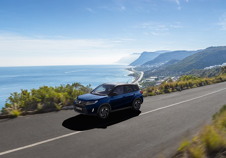 Pärchen läuft gemeinsam in Richtung eines Suzuki Vitara Hybrid in Blue Sphere Pearl Metallic.